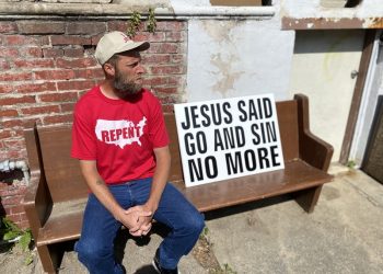 Damon Atkins sits on a bench outside his home in central Reading. A sign sits besides him with the words, "Jesus Said Go and Sin No More."
