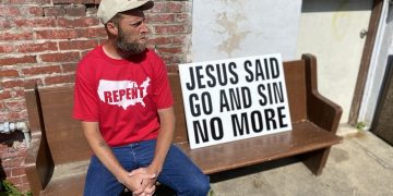 Damon Atkins sits on a bench outside his home in central Reading. A sign sits besides him with the words, "Jesus Said Go and Sin No More."